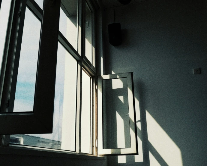 Sunlight streams through tall, modern windows with open glass panels, creating geometric shadows and a bright, airy interior filled with natural light.