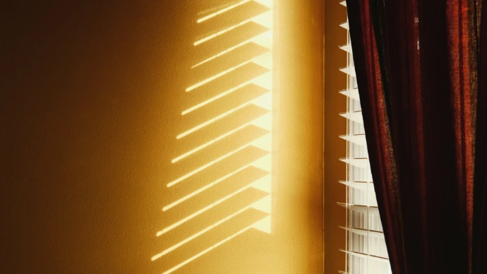 Sunlight streams through adjustable white horizontal window blinds, casting angled shadows on a warm interior wall and demonstrating precise light control.