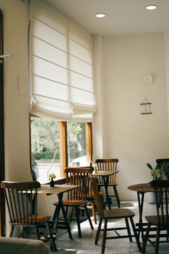 Custom white fabric Roman shades on tall cafe windows, featuring elegant folds that softly filter natural light and complement a warm, inviting interior.
